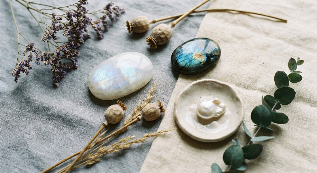 A softly lit flat lay of moonstone, labradorite, and pearl crystals arranged with dried botanicals and a small ceramic dish on a weathered linen surface.
