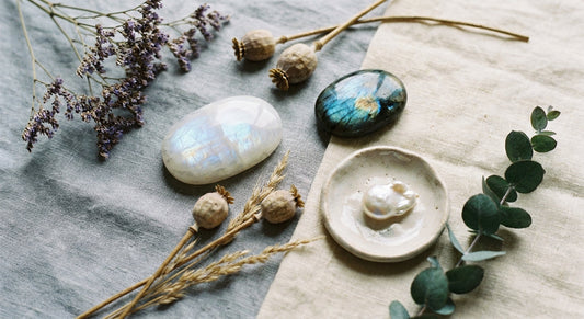 A softly lit flat lay of moonstone, labradorite, and pearl crystals arranged with dried botanicals and a small ceramic dish on a weathered linen surface.