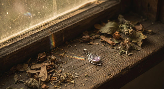 A delicate crystal pendant resting on a worn wooden surface beside dried botanicals, bathed in warm afternoon light with a soft prismatic glow.