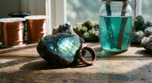 A raw labradorite stone and a partially electroformed copper ring rest on a weathered wooden studio surface, surrounded by copper wire and mineral specimens in warm, earthy light.