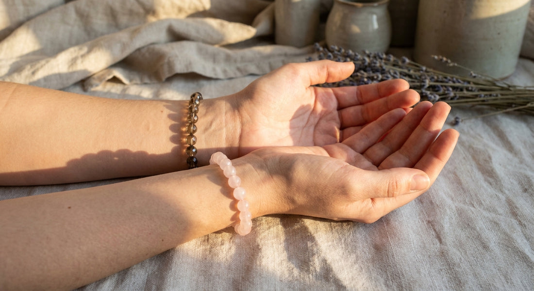 A woman's two wrists, each wearing a delicate crystal bracelet, rest softly on linen in warm natural light, evoking quiet ritual and energetic intention.