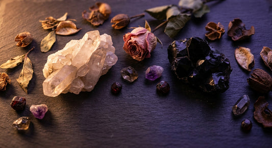 A moody flat lay of pale and dark crystals on slate stone, caught between warm golden and cool violet light, surrounded by dried botanicals and raw gemstones.