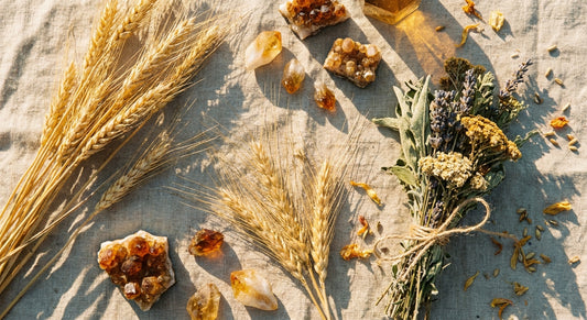 A golden flat lay of wheat stalks, amber crystals, and dried herb bundles on worn linen, bathed in warm late-summer light.