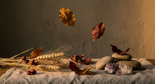 A sparse autumn still life of dried wheat, rosehips, fallen leaves, and raw crystals arranged on linen in warm golden light, with one leaf suspended mid-fall against a dark background.