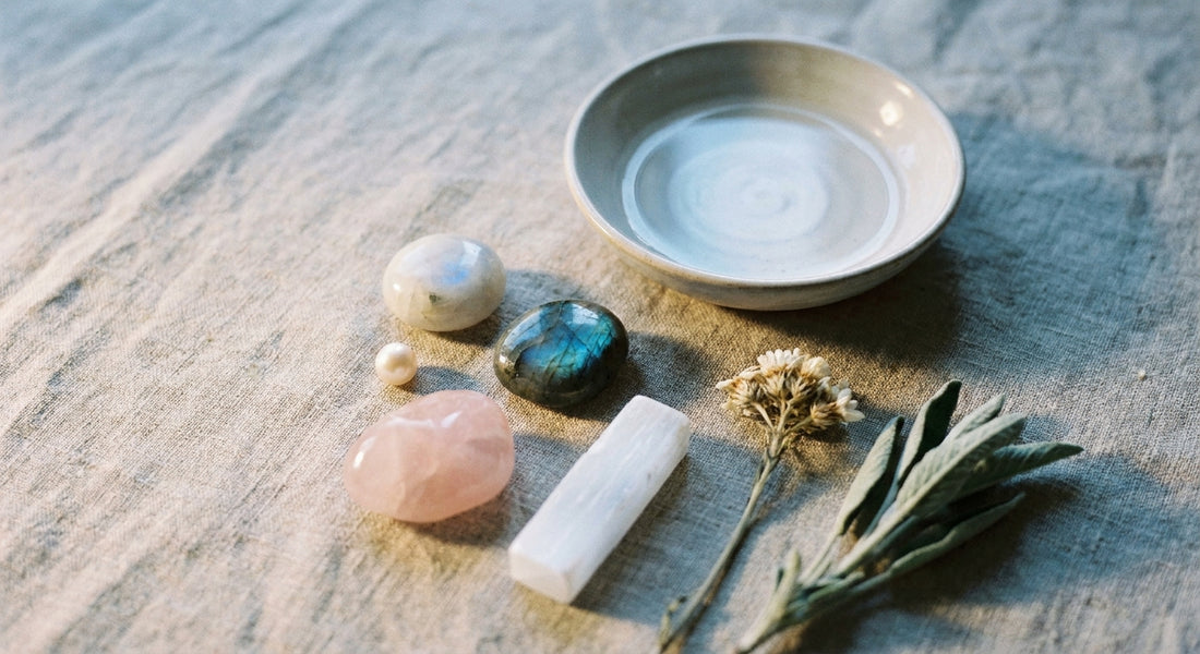 A softly lit flat lay of moonstone, pearl, labradorite, rose quartz, and selenite crystals arranged on weathered linen beside a small ceramic dish of water and a dried sprig of sage, evoking a quiet lunar ritual.