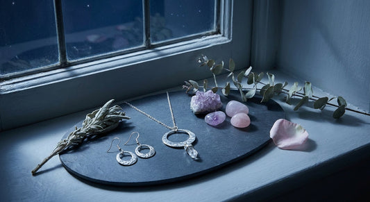 Delicate crystal-adorned jewelry arranged beside dried botanicals, rose quartz, and amethyst stones on a stone surface in cool moonlight, evoking a quiet ritual moment.
