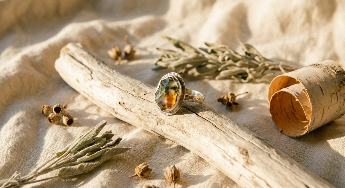 A single handcrafted gemstone ring resting on driftwood surrounded by dried botanicals and sage on a linen surface in warm golden light.