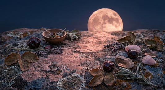 A glowing rose-gold full moon rises over a weathered stone surface arranged with raw crystals and dried botanicals in the quiet of a midsummer night.