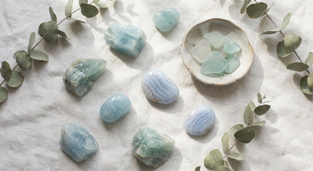 Raw aquamarine and blue lace agate crystals arranged on white linen with dried eucalyptus and a small ceramic dish, bathed in soft natural light.