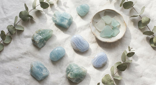 Raw aquamarine and blue lace agate crystals arranged on white linen with dried eucalyptus and a small ceramic dish, bathed in soft natural light.
