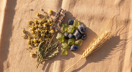 A sun-warmed flat lay of dried chamomile, lavender sprigs, raw peridot and sapphire crystals, and a wheat stalk arranged on aged linen in soft golden light.