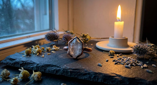 A handcrafted copper and crystal ring rests on dark slate surrounded by dried wildflowers and a glowing candle, bathed in soft moonlight.
