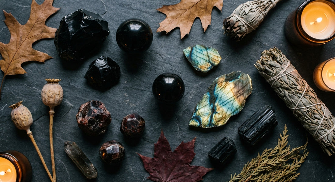 A moody flat lay of obsidian, labradorite, garnet, and black tourmaline crystals arranged with dried autumn botanicals on a dark slate surface, lit by warm candlelight.